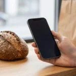 Dłonie właściciela piekarni z telefonem nad blatem z pieczywem, ciepłe poranne światło w sklepie
